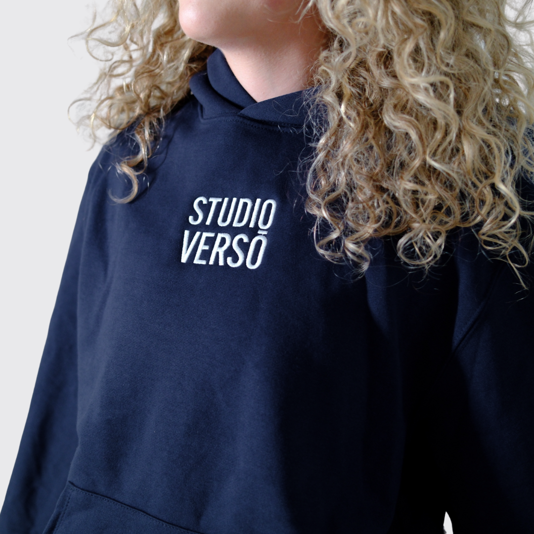 Navy hoodie with stacked Studio VERSŌ logo worn by a person with curly hair.