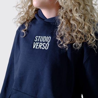 Navy hoodie with stacked Studio VERSŌ logo worn by a person with curly hair.